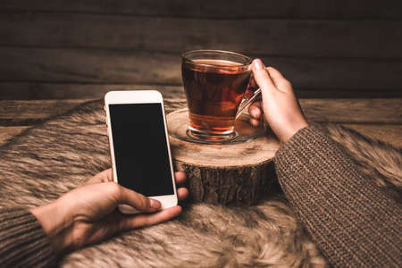 cup with tea and phone in the hands of a girl on a wooden backgroundの写真素材