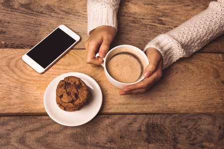white phone, a cup of coffee in the hands of the girl, cookies on the table on a wooden backgroundの写真素材