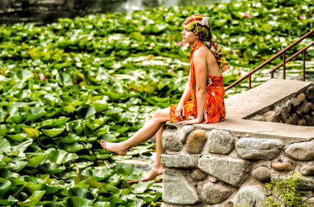 a woman is sitting on a stone pier looking at the lake with lotusの写真素材