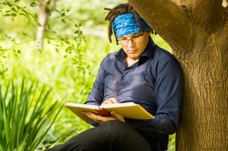 young man with dreadlocks reads a book in a parkの写真素材