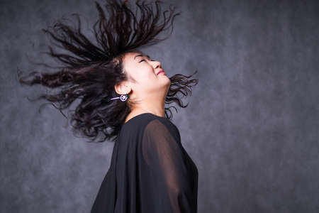attractive asian joyful happy female model is shaking head with brunette hair in studio wearing black dress, isolated on gray backgroundの写真素材