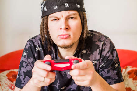 young man with dreadlocks and serious face playing video games with a joystick sitting on an orange couchの写真素材
