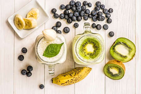 banana and kiwi milkshakes in mason jars with creme and decorations on top and decorated with blueberries, bananas, and kiwis. flat lay top view smoothie on wooden backgroundの写真素材