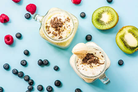 two banana and kiwi milkshakes in mason jars with creme and chocolate chips on top decorated with kiwis, raspberries and blueberries. flat lay top view smoothies on blue fashion backgroundの写真素材