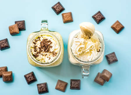 two banana and kiwi milkshakes in mason jars with creme on top decorated with chocolate pieces. flat lay top view smoothie on blue fashion background. creative layout with chocolate and milkshakesの写真素材