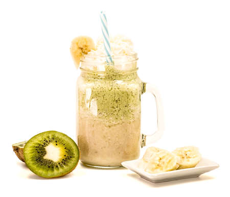banana and kiwi milkshake in mason jar with creme on top and drinking straw decorated with slices of fruit isolated on whiteの写真素材