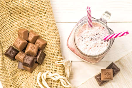 berry and fruit milkshake top view on white wooden background decorated with pieces of chocolate and coffee bag. summer drinks and desserts. fresh natural organic milkshake with two drinking straws flat layの写真素材