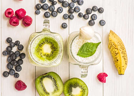 banana and kiwi milkshakes in mason jars with creme and decorations on top and decorated with blueberries, raspberries, bananas, and kiwis. flat lay top view smoothie on wooden backgroundの写真素材