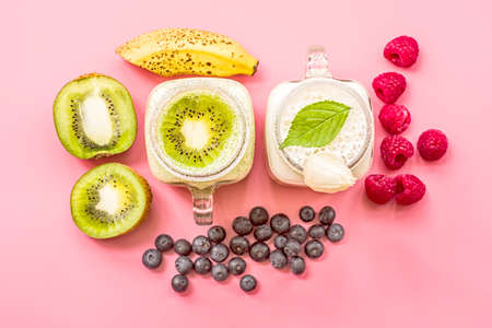 two banana and kiwi milkshakes in mason jars with creme on top decorated with kiwis, bananas, raspberries and blueberries. flat lay top view smoothie on pinkの写真素材