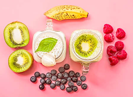 two banana and kiwi milkshakes in mason jars with creme on top decorated with kiwis, bananas, raspberries and blueberries. flat lay top view smoothie on pinkの写真素材