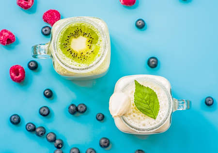 two banana and kiwi milkshakes in mason jars with creme on top decorated with raspberries and blueberries. flat lay top view smoothie on blue backgroundの写真素材