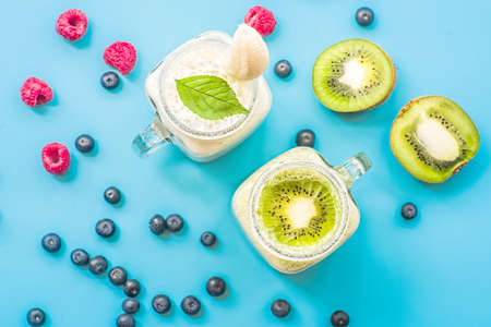 two banana and kiwi milkshakes in mason jars with creme on top decorated with raspberries and blueberries. flat lay top view smoothie on blueの写真素材