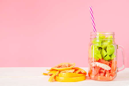 cut fresh fruit in a mason jar with a drinking straw and grapefruit peel on pink and white woodの写真素材