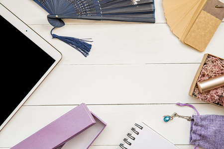 top view flat lay of workspace with a tablet, fan, lipstick, paper tags and decorations on white...