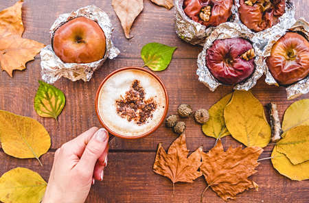 hand with a cup of coffee and backed apples in foil with autumn leaves on wooden backgroundの写真素材