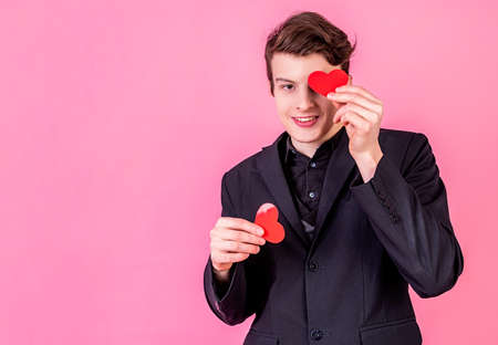 Valentines Day concept. Young smiling man holding heart shaped valentine cards in front of his eyes on pink background with copy spaceの写真素材