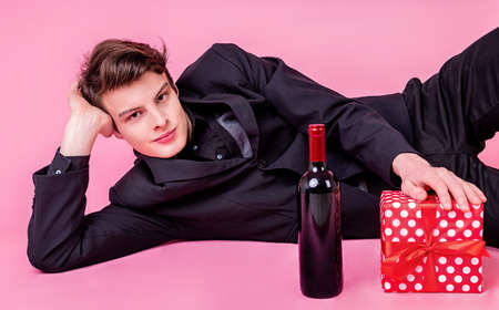 Valentines day concept. Young stylish man laying on the pink background with a red gift box and a wine bottleの写真素材