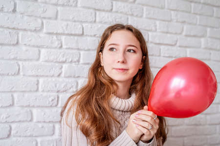 Valentines Day concept. Young woman holding a red balloon on white brick wall background