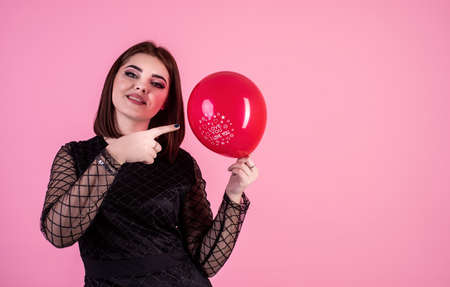 woman in black dress pointing at the red balloon isolated on pink background with copy spaceの写真素材