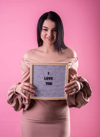 Beautiful woman in beige dress holding a felt letter board with the words I Love You on pink backgroundの写真素材