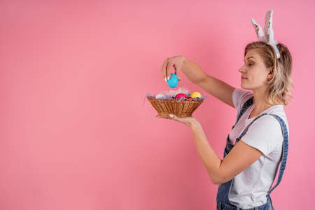 Easter holiday concept. Cheerful young woman with bunny ears putting colored egg into the basket isolated on pink background with copy spaceの写真素材