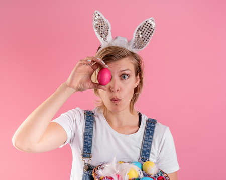 Easter concept. Surprised young woman in bunny ears holding colored chicken eggs in front of her eye...