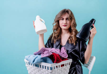 Laundry concept. puzzled housewife holding a basket of clothes and detergent bottles ready for laundry isolated on blue background with copy spaceの写真素材