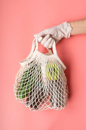Coronavirus food supplies concept. Food delivery. Hand in a glove holding white eco friendly mesh bag with food supplies on pink backgroundの写真素材