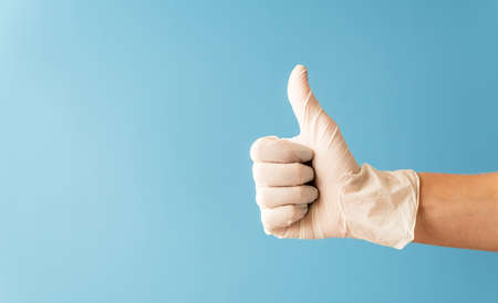 Hand wearing white rubber glove giving the thumbs up signal on blue backgroundの写真素材