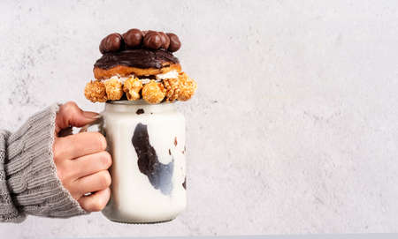 Female hand holding ice-cold chocolate donut milkshake in a mason jar decorated with caramel popcorn, whipped cream and cocoa candies front view with copy spaceの写真素材