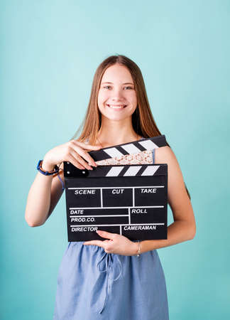 Happy smiling teenager girl in a blue dress holding a clapper board isolated on blue backgroundの写真素材