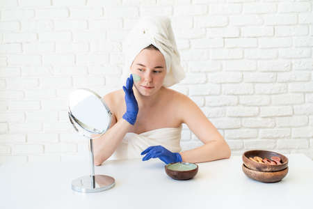 Spa and wellness. Natural cosmetics. Self care. Close up of happy young caucasian woman in white bath towels wearing gloves applying clay face mask looking at the mirrorの写真素材