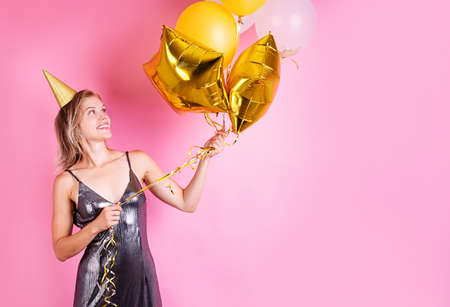 Young blond woman wearing birthday hat holding golden balloons celebrating birthday on pink...
