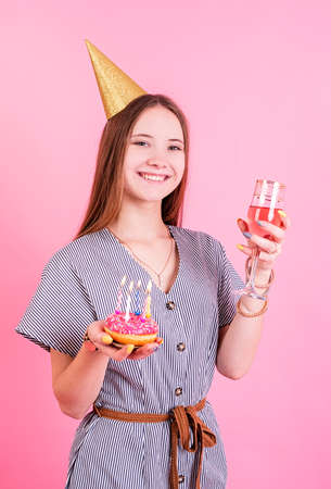 Smiling Teenager birthday girl in birthday hat holding two donuts with candles isolated on pink