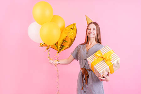 Young woman in a birthday hat holding balloons and big gift box celebrating birthday party isolated...