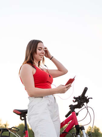 Teenage girl standing next to her bike listening to the music in headphones in the park at sunset....