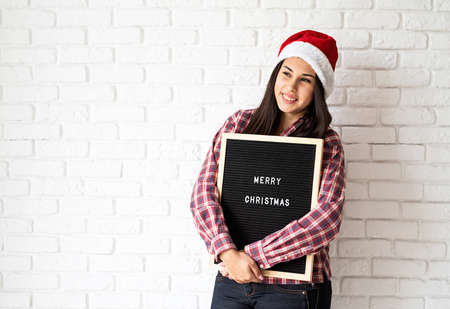Portrait of a happy beautiful latin woman in santa hat with black letter board with the words Merry Christmas on white brick wall background with copy spaceの写真素材