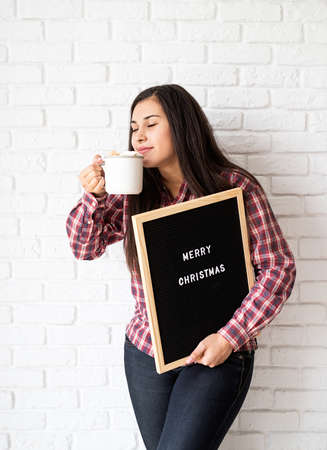 Portrait of a happy beautiful latin woman with letter board with the words Merry Christmas holding a cup of cocoa with marshmallowsの写真素材