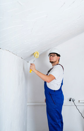 Repair, home improvement and renovation concept. Closeup of a worker in blue overalls with a roller for coloring walls on white wall background with copy spaceの写真素材