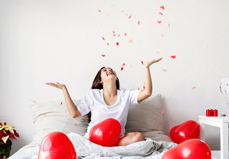 Valentines Day. Young happy brunette woman laying in the bed with red heart shaped balloons and decorationsの写真素材