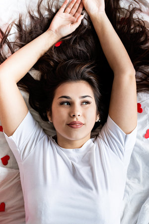 Valentines Day. Young brunette woman sitting awake in the bed with red heart shaped balloons and decorations drinking coffee eating croissantsの写真素材