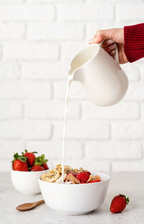Healthy eating and dieting. Female hand pouring milk to breakfast cereal bowl on white brick wall backgroundの写真素材