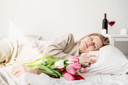Happy smiling woman sitting on the bed wearing pajamas, with pleasure enjoying flowers and reading a...
