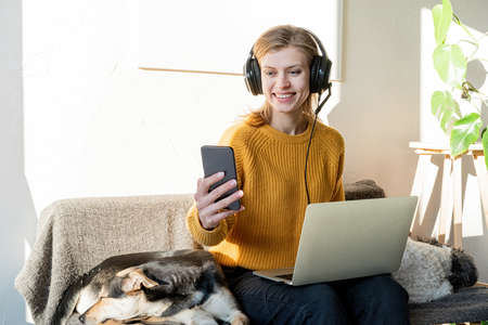 Distance learning. E-learning. Young smiling woman in yellow sweater and black headphones studying...