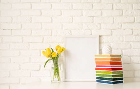 Bouquet of yellow tulips in a glass vase, pile of colorful books and blank photo frame on a white brick wall background. Mock up designの写真素材
