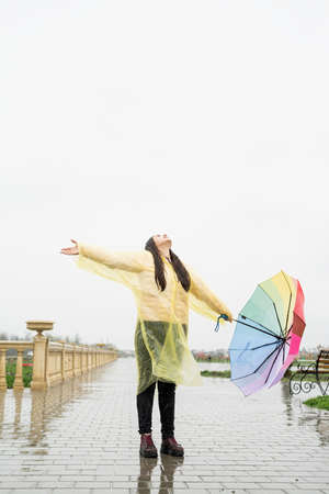 Beautiful brunette woman in yellow raincoat holding rainbow umbrella out in the rain