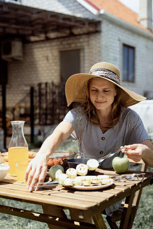 Young woman in summer hat grilling meat outdoors in the backyard, sitting with her dog, giving pet a snackの写真素材