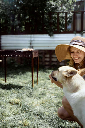 Young woman in summer hat grilling meat outdoors in the backyard, sitting with her dog, giving pet a snackの写真素材