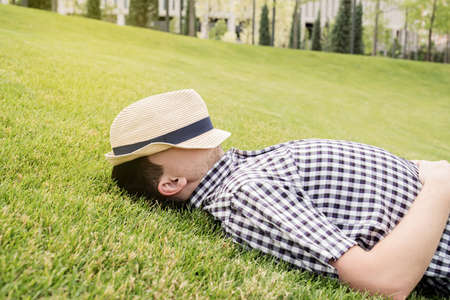 Young thoughtful man in summer hat sitting on the grass in the park, looking awayの写真素材