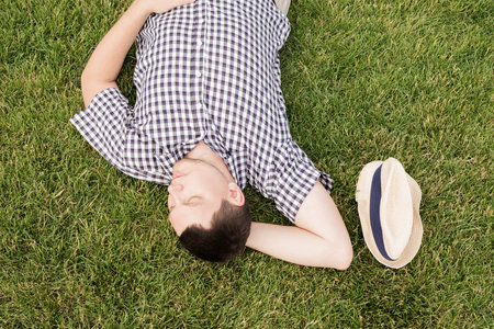 Young thoughtful man in summer hat sitting on the grass in the park, looking awayの写真素材